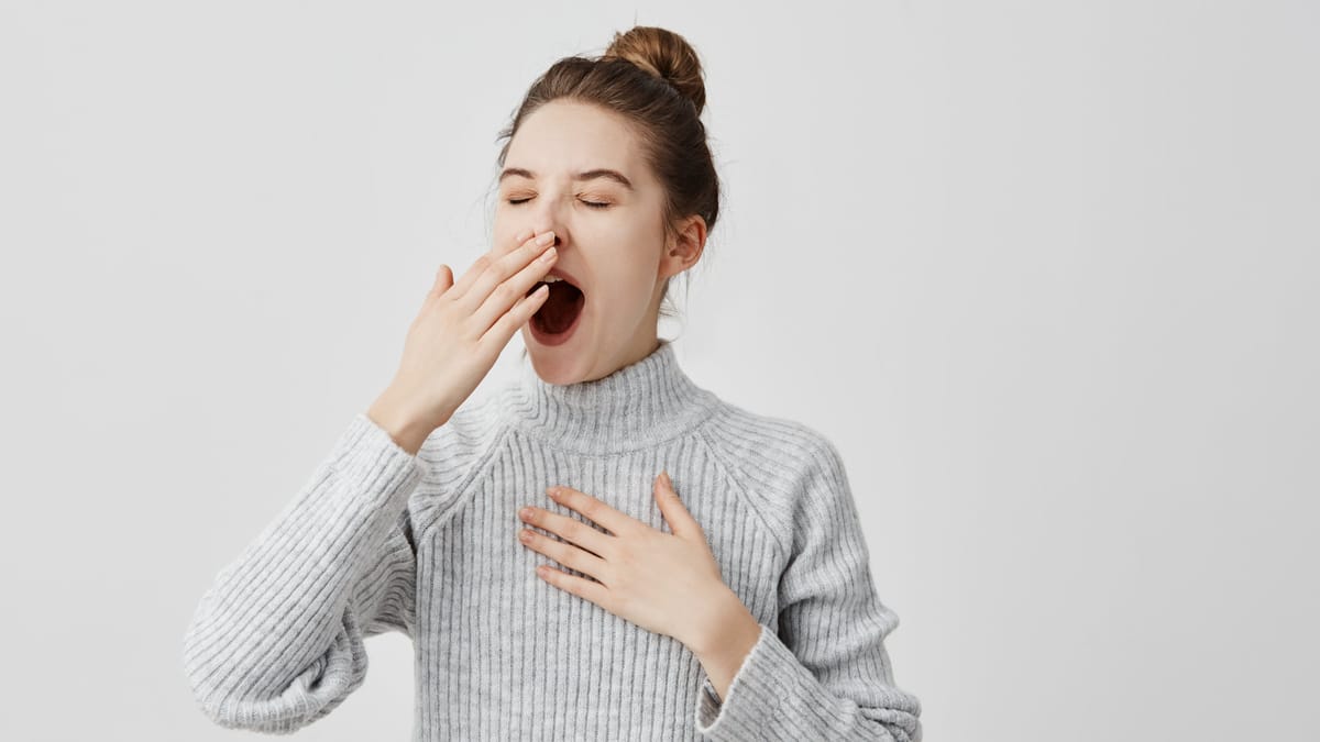 Junge Frau im grauen Rollkragenpullover gähnt mit geschlossenen Augen und hält sich höflich eine Hand vor den Mund, die andere ruht auf der Brust.