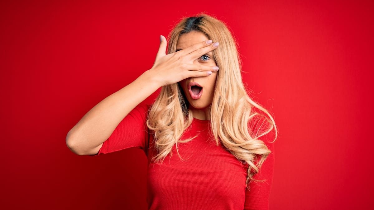 Junge Frau mit langen blonden Haaren hält sich eine Hand vor das Gesicht und schaut überrascht durch die Finger.