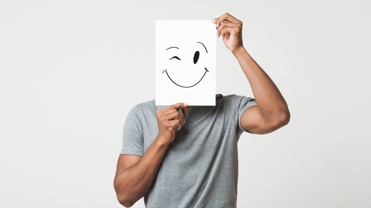 Eine Mann in grauem T-Shirt hält ein weißes Blatt Papier vor ihr Gesicht, auf dem ein zwinkerndes Smiley-Gesicht gezeichnet ist.