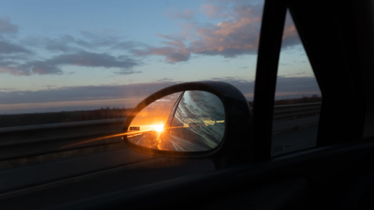 Seitenspiegel eines fahrenden Autos auf der Autobahn, in dem sich ein goldener Sonnenuntergang spiegelt. Pastellfarbener Abendhimmel im Hintergrund.