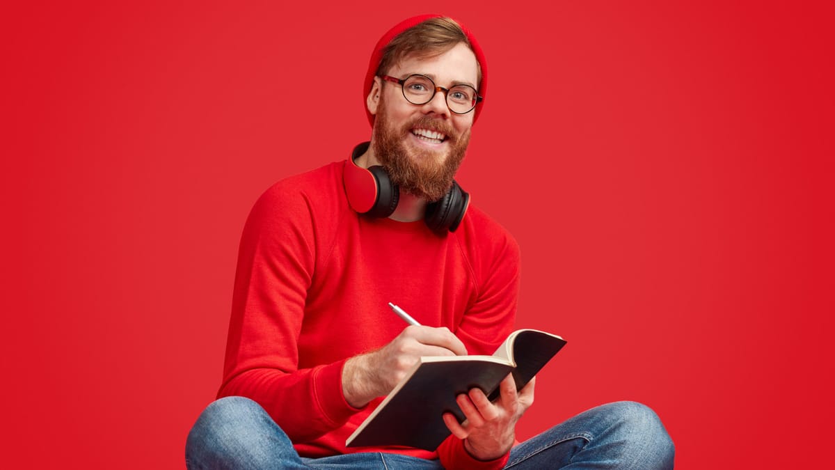 Junger Mann mit roter Mütze, Brille und Kopfhörern um den Hals sitzt im Schneidersitz und schreibt lächelnd in ein Notizbuch.