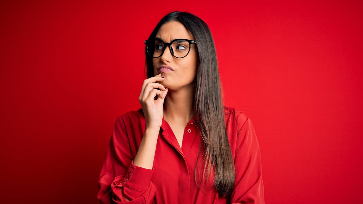 Junge Frau mit schwarzer Brille und roter Bluse vor rotem Hintergrund, Hand nachdenklich ans Kinn gelegt, Blick zur Seite gerichtet.
