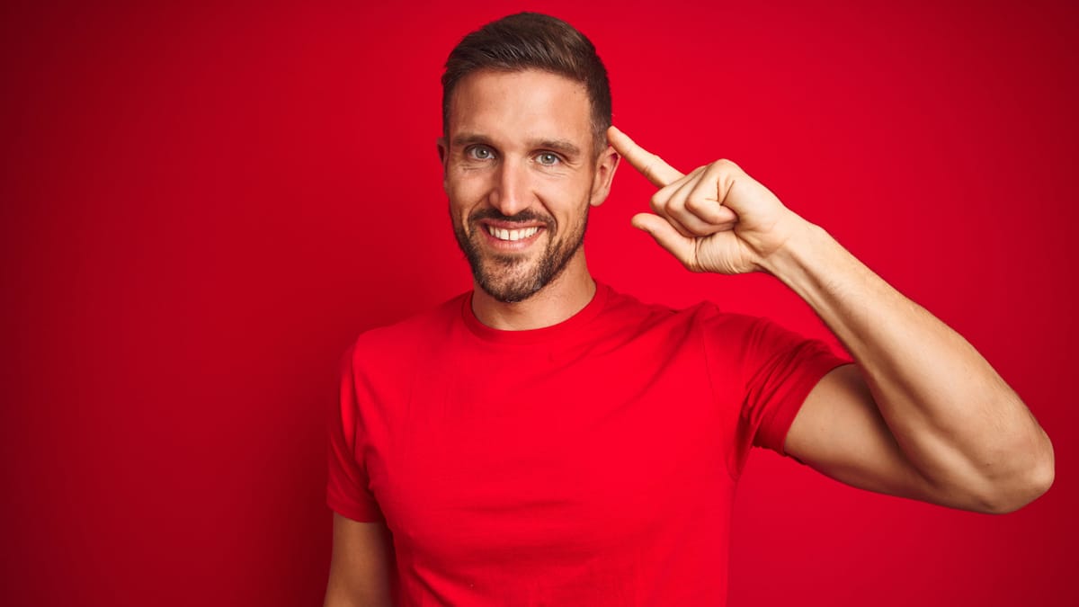 Lächelnder Mann mit Bart im roten T-Shirt vor rotem Hintergrund, Zeigefinger an die Schläfe gelegt als Geste für "Denken" oder "gute Idee"