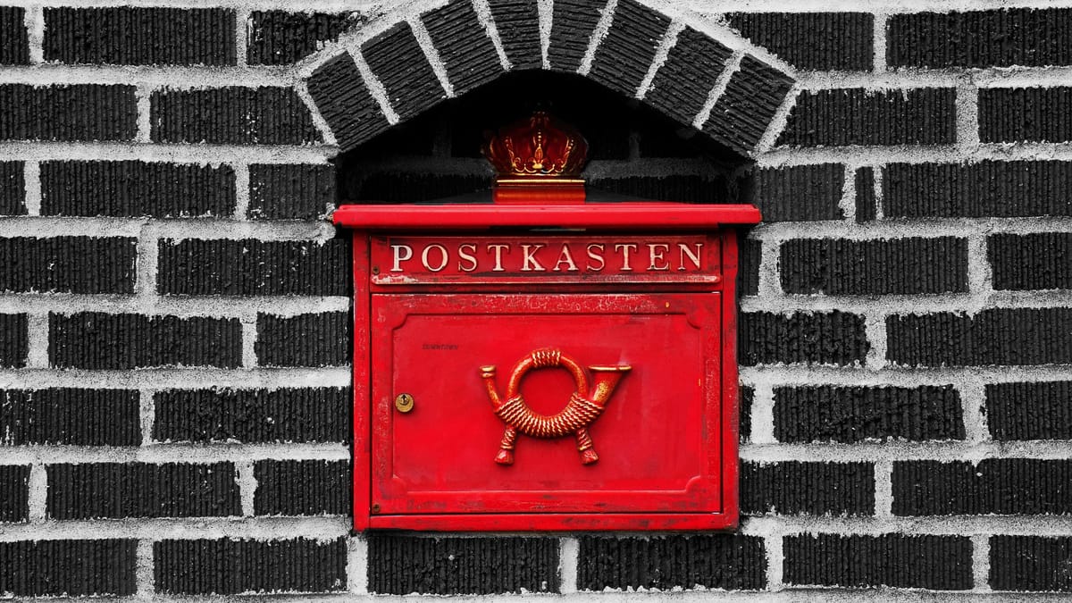 Roter historischer Briefkasten mit Aufschrift "POSTKASTEN" und goldenem Posthorn, eingemauert in schwarz-weißer Backsteinwand unter einem Rundbogen mit goldener Krone.
