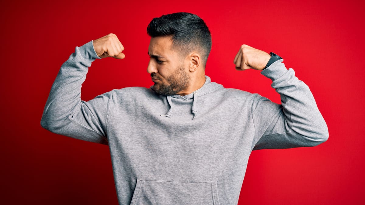 Mann mit Bart in grauem Hoodie steht vor rotem Hintergrund und flexed beide Arme mit entschlossenem Blick zur Seite.