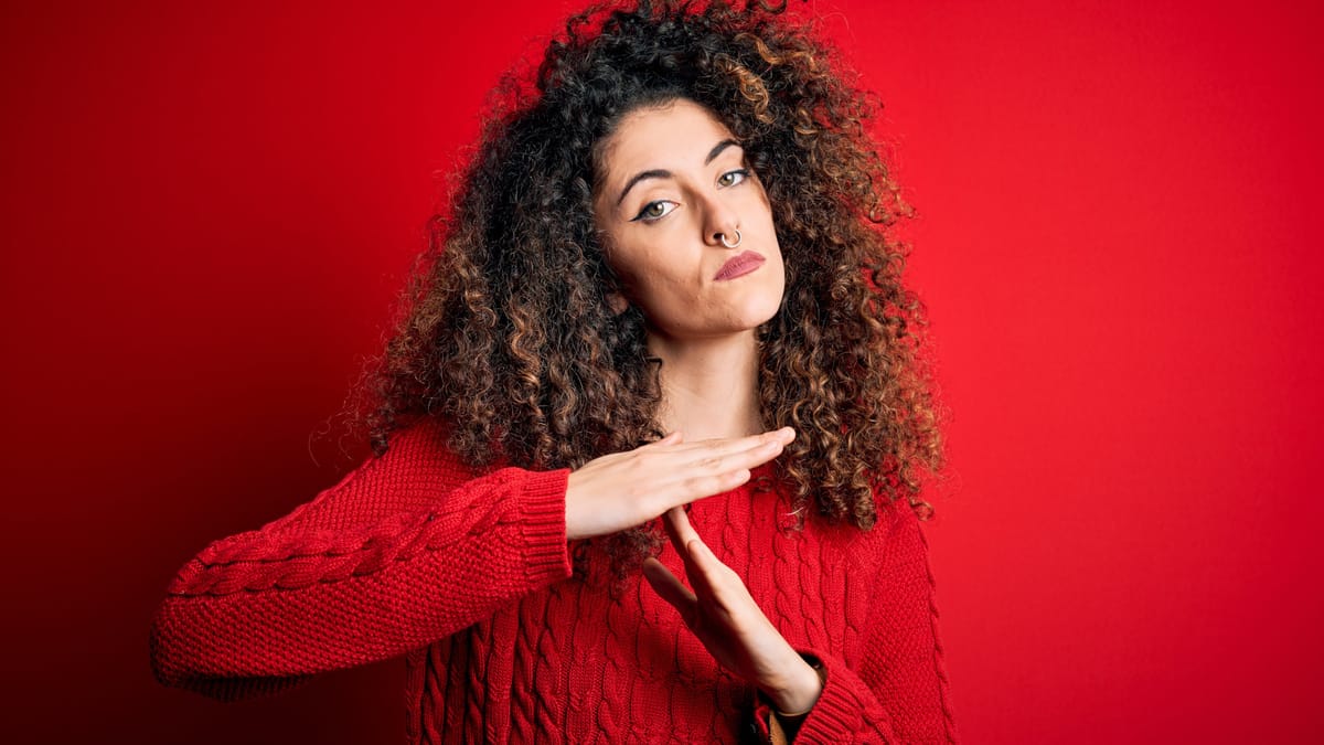Frau mit lockigem Haar und rotem Pullover macht vor einem roten Hintergrund die Time-out-Geste.
