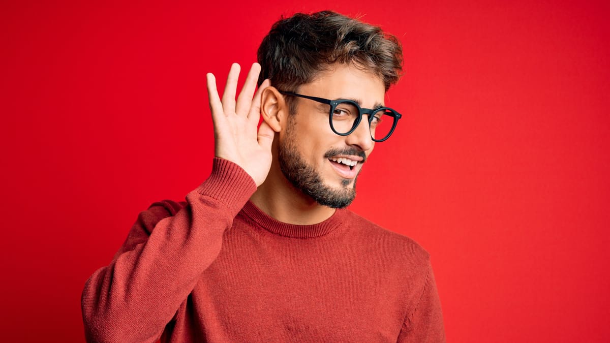 Mann mit Bart und Brille in rotem Pullover steht vor rotem Hintergrund und hält sich lächelnd die Hand ans Ohr als würde er lauschen.