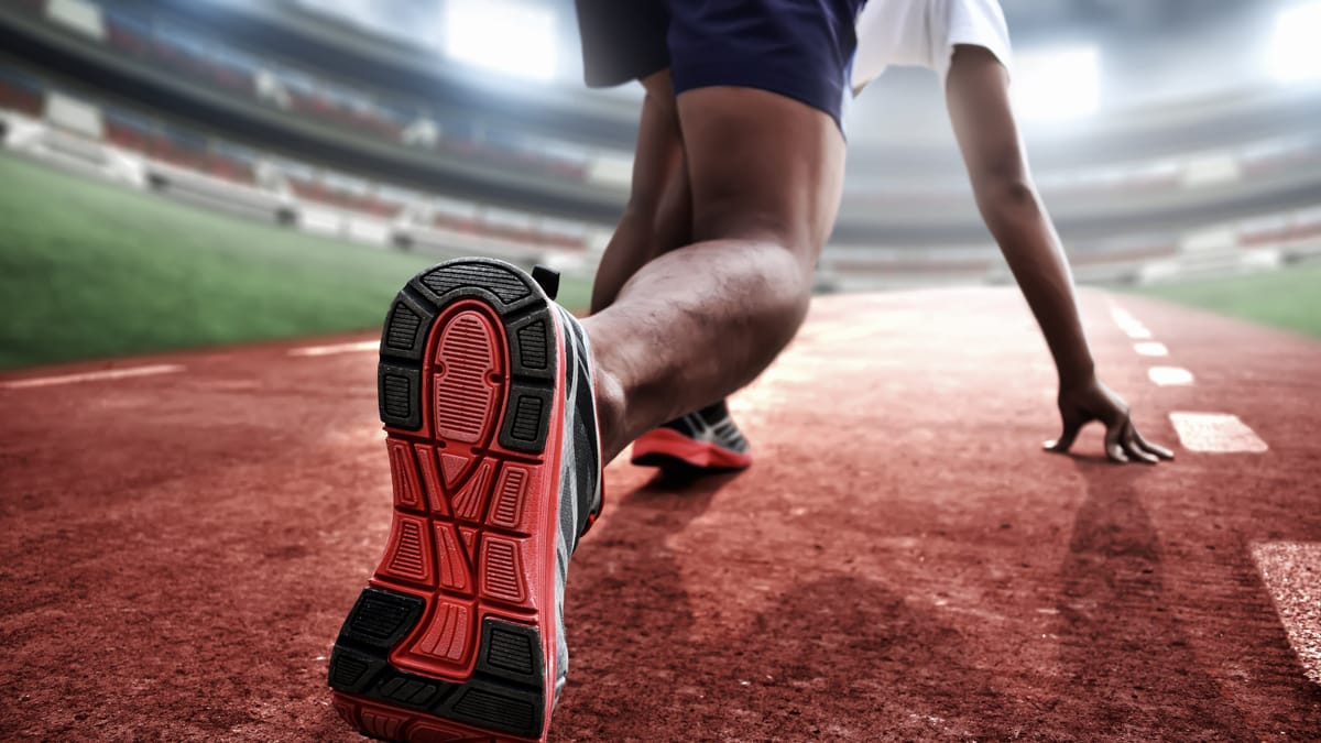 Sprinter kauert in der Startposition auf einer roten Laufbahn in einem beleuchteten Stadion, die Schuhsohle der Nahaufnahme deutlich im Vordergrund.