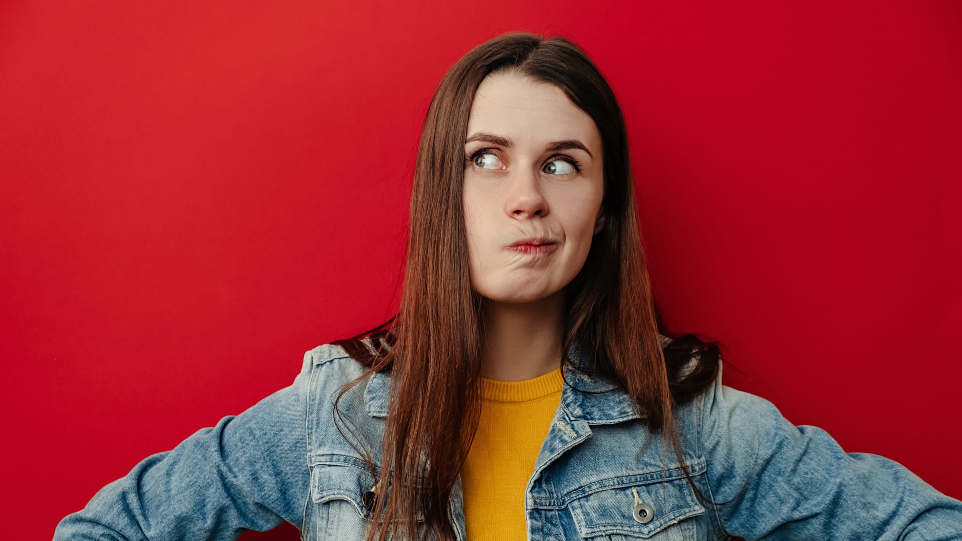 Junge Frau mit langen braunen Haaren steht vor einer roten Wand, schaut nachdenklich zur Seite und zieht die Lippen zusammen, als würde sie überlegen oder zögern.
