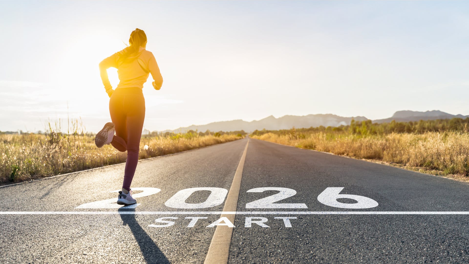 Eine Joggerin läuft auf einer Straße dem Horizont entgegen. Über der Startlinie steht in großen weißen Zahlen 2026.