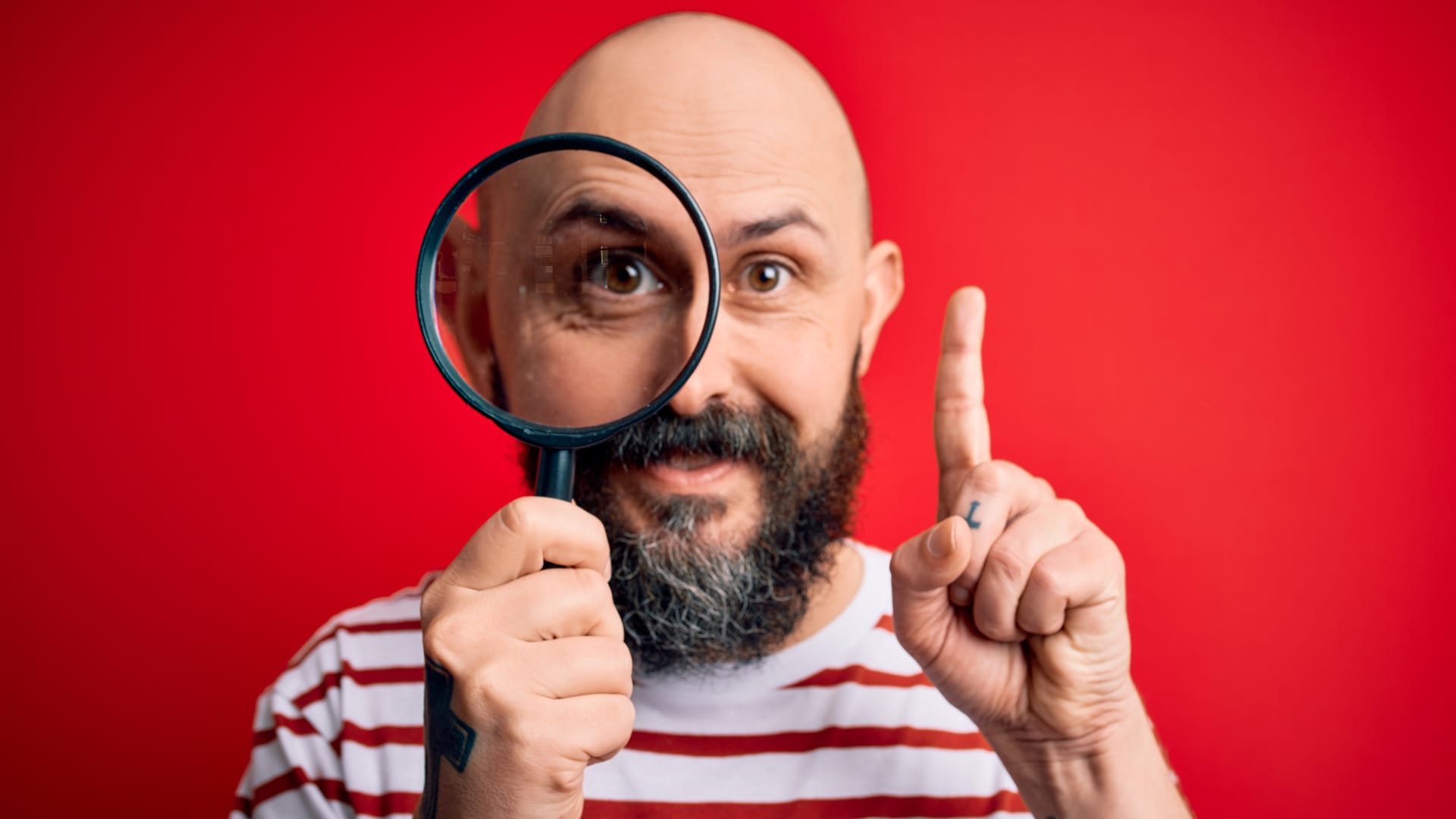 Kahlköpfiger Mann in rot-weiß gestreiftem Shirt, der eine Lupe vor sein rechtes Auge hält und den Zeigefinger der rechten Hand nach oben reckt.